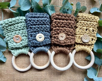 Puede incluir: Cuatro anillos de dentición para bebés hechos a mano con ganchillo en tonos verdes, azules, marrones y amarillos. Cada anillo presenta un anillo de madera y un cierre de botón. Los artículos se exhiben sobre una superficie de arpillera natural con hojas de eucalipto.