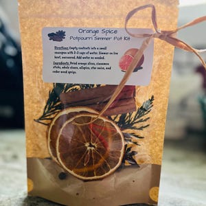 以下が含まれることがあります： 「Orange Spice Potpourri Simmer Pot Kit」とラベルされた茶色の紙袋で、ラフィアで結ばれています。 袋には、乾燥オレンジスライス、シナモンスティック、その他のスパイスが入っています。 ラベルには指示と材料が含まれています。