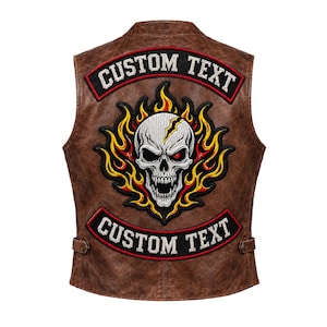 Può includere: Un gilet in pelle marrone con una grande toppa ricamata sul retro. La toppa mostra un teschio bianco con una crepa, circondato da fiamme gialle e arancioni. Due fasce nere curve riportano il testo bianco CUSTOM TEXT sopra e sotto il disegno.