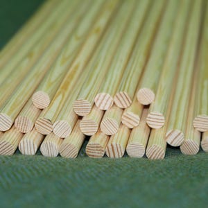 Puede incluir: Una colección de varillas de madera de color claro dispuestas en un haz paralelo. Cada varilla cilíndrica presenta un patrón de veta natural visible con tonos bronceados y crema, mostrando la sección transversal circular del extremo.
