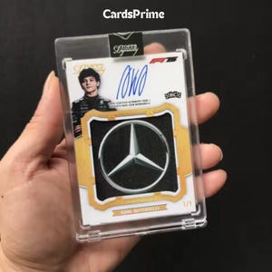 May include: A Topps Dynasty Formula 1 trading card featuring a blue ink autograph of Kimi Antonelli, a portrait of the driver, and a silver Mercedes-Benz logo emblem embedded in the card. The card is encased in a clear protective plastic holder.
