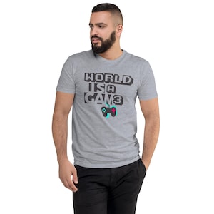 May include: A heather gray short-sleeve t-shirt featuring the pixelated black text "WORLD IS A GAM3" on the chest. Below the text is a graphic of a black video game controller with a teal cord and a small pink button detail on the right side.