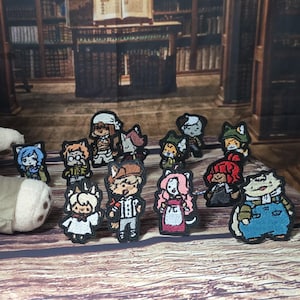 May include: A plush cat doll wearing a white shirt and tan overalls sits beside a collection of small, embroidered patches featuring various cartoon characters with distinct outfits, hairstyles, and colours, arranged on a wooden surface.
