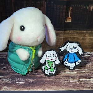 May include: A white plush rabbit toy wearing a teal puffer vest with a yellow zip, accompanied by two embroidered patches featuring cartoon rabbits, one wearing a green outfit and the other wearing a blue jumper, both with black borders.