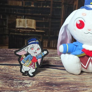 Puede incluir: Un parche bordado que muestra un conejo blanco con sombrero azul, chaqueta azul y chaleco rojo, junto a un peluche del mismo personaje con ojos rojos, sombrero azul y chaqueta azul con ribetes amarillos.
