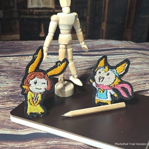 Puede incluir: Dos parches bordados con personajes de dibujos animados con orejas amarillas, colocados cerca de un maniquí de madera y un pequeño lápiz de madera. Un parche muestra un personaje con traje amarillo y el otro un personaje con bufanda rosa.
