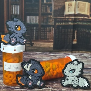 Pode incluir: Três emblemas bordados com criaturas estilizadas de olhos grandes. Um emblema cinza-escuro com detalhes laranja está sobre um frasco de comprimidos de plástico laranja com o rótulo Mind Wipe. Dois emblemas adicionais, um cinza-escuro e um cinza-claro, estão próximos.