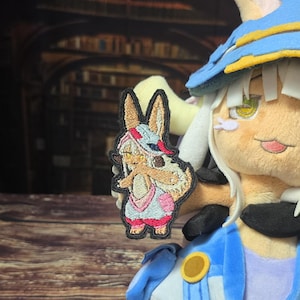 Peut inclure: Un écusson brodé représentant un personnage beige aux longues oreilles, portant un chapeau bleu et blanc ainsi qu'une tenue rose et blanche, tenu devant une peluche assortie coiffée d'un chapeau bleu avec de grands yeux jaunes.