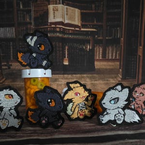 May include: A collection of six embroidered patches featuring stylized chibi-style creatures in various colors including black, white, yellow, and pink, each with distinct eye colors and small paws, arranged in a group on a wooden surface.