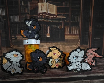 Può includere: Una collezione di sei toppe ricamate raffiguranti creature in stile chibi in vari colori, tra cui nero, bianco, giallo e rosa, ognuna con colori degli occhi distinti e piccole zampe, disposte in gruppo su una superficie di legno.