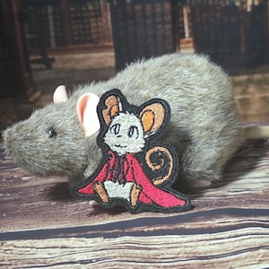 以下が含まれることがあります： 大きな耳を持つ漫画のネズミのキャラクターが描かれた刺繍ワッペン。白い体に赤いマントを羽織っています。ワッペンには黒いステッチの縁取りがあり、ピンクの耳を持つリアルな灰色のネズミのぬいぐるみの前に配置されています。