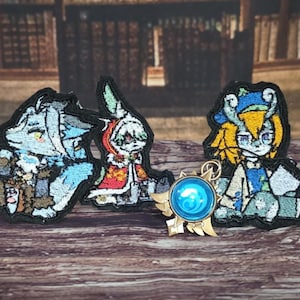 Puede incluir: Tres parches bordados con personajes de estilo anime y un dije metálico circular con un centro azul. Los parches muestran un trabajo de hilo detallado en tonos azul, rojo, amarillo y blanco, mientras que el dije tiene detalles en tono dorado.