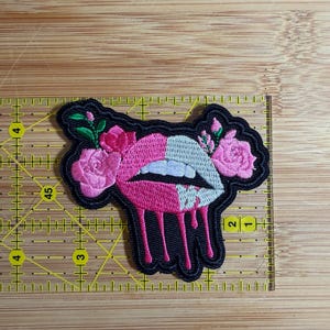 May include: An embroidered patch featuring a split pink and white lip design with pink roses and green leaves. The lips appear to be dripping pink liquid. A yellow ruler is positioned behind the patch, showing it is approximately 3.5 inches wide.