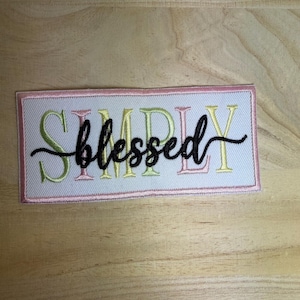 May include: A rectangular embroidered patch with a white background and a pink border. The word SIMPLY is stitched in alternating light green and pink letters, with the word blessed embroidered in black cursive script across the center.