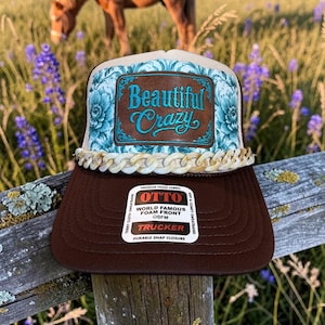 May include: A brown trucker hat featuring a teal and white floral patterned front panel with a brown leather patch embroidered with the text Beautiful Crazy. The hat includes a decorative beige chain trim and a white Otto brand sticker on the brim.