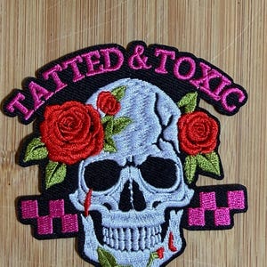 May include: An embroidered patch featuring a light blue skull with red roses and green leaves. The text TATTED & TOXIC is stitched in pink above the skull. The design includes pink checkered accents on a black background with red details.