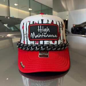May include: A trucker hat with a red brim and a white front panel featuring black vertical stripes and red roses. A black rectangular patch reads High Maintenance in white script. A black chain with rhinestones and three silver studs adorn the brim.