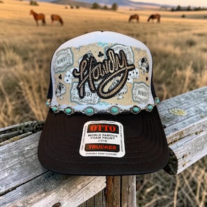 May include: A black and white trucker hat featuring a rope-style Howdy script embroidery on the front panel. The design includes playing card motifs, a turquoise stone trim, and a sticker on the brim reading Otto World Famous Foam Front.