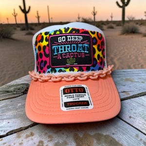 Puede incluir: Una gorra de camionero con visera color melocotón y frente de espuma blanca con un patrón de leopardo colorido. Un parche rectangular negro muestra el texto GO DEEP THROAT A CACTUS con un icono de cactus. Una cadena de plástico melocotón adorna la base.