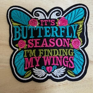 Puede incluir: Un parche bordado negro con forma de mariposa, alas color turquesa, rosas fucsias y hojas verdes. El texto dice: IT'S BUTTERFLY SEASON I'M FINDING MY WINGS. El diseño incluye remolinos decorativos blancos y un pequeño corazón rosa con alas blancas.