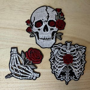 May include: Three embroidered patches featuring a white skull with red roses, a skeletal hand holding a red rose, and a human rib cage with a central red rose. Each patch is detailed with black stitching and white thread on a fabric base.