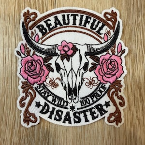 May include: An embroidered patch featuring a white steer skull with black horns, adorned with pink roses and brown scrollwork. The text reads BEAUTIFUL at the top and STAY WILD 100 PROOF DISASTER at the bottom in black lettering on a white background.