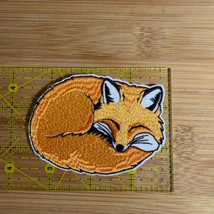 Op de afbeelding: Een geborduurde patch van een opgerolde oranje vos met witte accenten en zwarte details. De patch ligt op een gele liniaal en meet ongeveer 8,9 cm in de breedte en 5,7 cm in de hoogte.