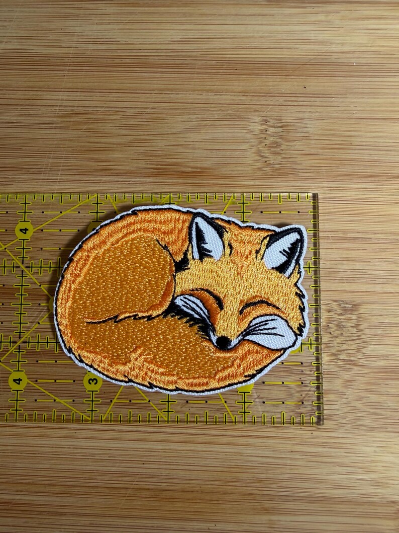 Op de afbeelding: Een geborduurde patch van een opgerolde oranje vos met witte accenten en zwarte details. De patch ligt op een gele liniaal en meet ongeveer 8,9 cm in de breedte en 5,7 cm in de hoogte.
