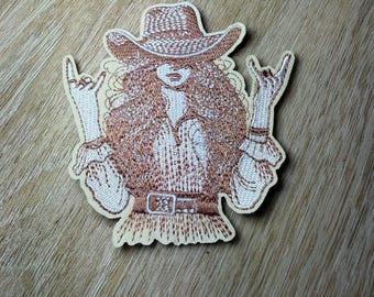 May include: An embroidered patch featuring a figure wearing a cowboy hat and a long-sleeved shirt with a belt. The figure has long hair and is making a rock and roll hand gesture with both hands. The design uses shades of brown and white thread.