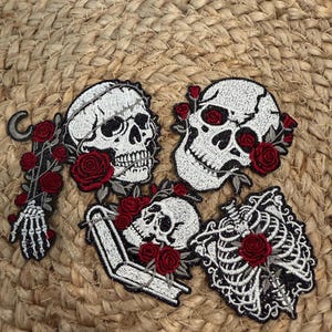 May include: Four embroidered patches featuring white skulls, a skeletal hand, and a rib cage, all accented with deep red roses. One patch includes a crescent moon and another features an open book. The designs use black, white, and red thread.