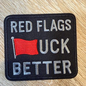 May include: A rectangular black embroidered patch with a stitched border. The text reads RED FLAGS in grey, followed by a red flag icon replacing the letter F, then UCK in grey, and the word BETTER in grey on the bottom line of the patch.