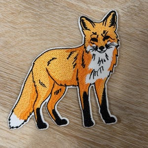 May include: An embroidered patch featuring a standing fox with vibrant orange fur, a white chest and tail tip, and black accents on the ears, legs, and facial features. The design is outlined in white thread, showcasing detailed stitching throughout the body.