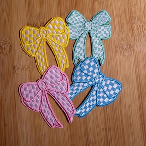 May include: Four embroidered patches in the shape of bows, each featuring a checkered pattern. The patches are colored in yellow, light green, pink, and blue, with each bow having a matching solid border around the edges.