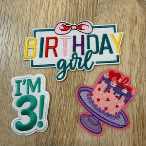 Peut inclure: Trois écussons brodés comprenant un écusson rectangulaire blanc avec un nœud rose et le texte BIRTHDAY girl en lettres multicolores, un écusson blanc avec le texte vert I'M 3!, et un gâteau rose sur un support violet avec des cœurs rouges.