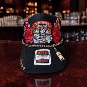 May include: A black trucker hat featuring an embroidered patch with red roses, flames, and the text I'D RATHER BE SOMEONE'S SHOT OF VODKA THAN EVERYONE'S CUP OF TEA. A gold chain adorns the brim with a miniature Tito's vodka bottle charm attached.