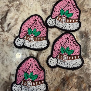 Könnte beinhalten: Vier gestickte Aufnäher in Form von Weihnachtsmannmützen in Rosa mit weißem Rand und Bommel. Jede Mütze hat ein braunes, gürtelartiges Band mit einem weißen, runden Akzent in der Mitte sowie grünen Stechpalmenblättern mit kleinen roten Beeren.