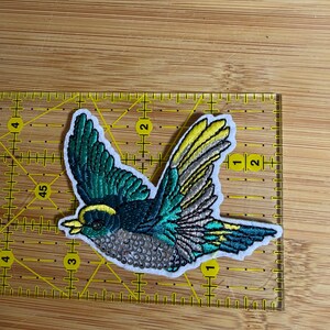May include: An embroidered bird patch featuring teal, yellow, and silver thread with a white border. The patch is positioned on a yellow ruler, showing a width of approximately 3.5 inches and a height of approximately 2.5 inches.
