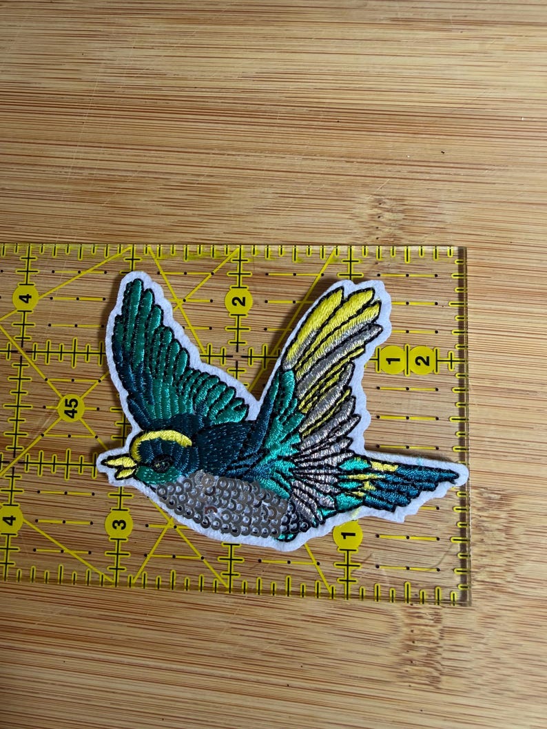 May include: An embroidered bird patch featuring teal, yellow, and silver thread with a white border. The patch is positioned on a yellow ruler, showing a width of approximately 3.5 inches and a height of approximately 2.5 inches.