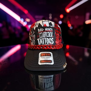 Pode incluir: Um boné trucker preto com um emblema frontal com o texto BAD NEWS: MORE TATTOOS, rodeado por gráficos de cartas de jogar e rosas. O boné inclui um detalhe de corrente de plástico vermelha na aba e um autocolante da marca Otto.