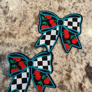 May include: Two embroidered bow-shaped patches featuring a black and white checkered pattern combined with a red cherry print on a black background. Each bow is outlined with a bright turquoise border and has a solid black center knot detail.