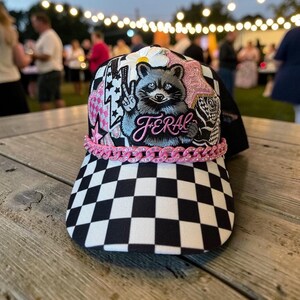 Könnte beinhalten: Eine schwarz-weiß karierte Baseballkappe mit einer zentralen Grafik eines Waschbären, der ein Friedenszeichen zeigt. Das Design umfasst rosa Sterne und Blitze, eine weiße Blume und das Wort FERAL in rosa Schrift sowie eine rosa Kettengarnitur.