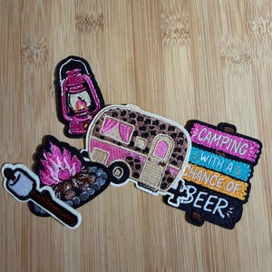 May include: Four embroidered patches featuring a pink lantern, a campfire with a marshmallow on a stick, a leopard print camper with pink accents, and a sign that reads CAMPING WITH A CHANCE OF BEER in pink, blue, orange, and white text.