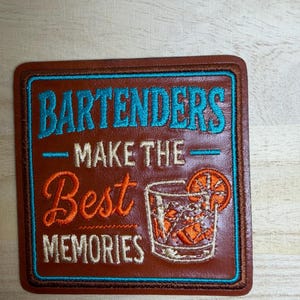 Puede incluir: Un parche cuadrado de cuero marrón con texto bordado que dice BARTENDERS MAKE THE Best MEMORIES en hilo azul, blanco y naranja. Incluye una ilustración bordada de un vaso con una rodaja de naranja como decoración en el parche.