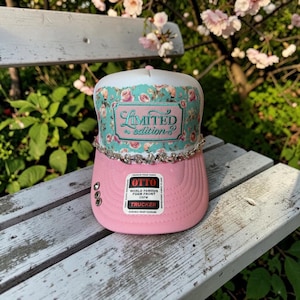 May include: A trucker hat with a pink brim and a white foam front panel featuring a teal floral pattern. The front displays the text Limited edition in cursive script. A row of clear rhinestones accents the base of the front panel.