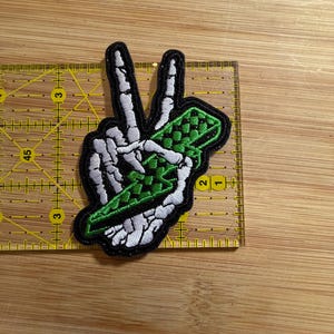 May include: An embroidered patch featuring a white skeleton hand making a peace sign while holding a green and black checkered lighter. The patch is positioned on a yellow ruler showing a width of approximately 2.5 inches.