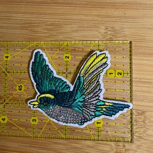 May include: An embroidered bird patch featuring teal, yellow, and silver thread with a white border. The patch is positioned on a yellow ruler, showing a width of approximately 3.5 inches and a height of approximately 2.5 inches.