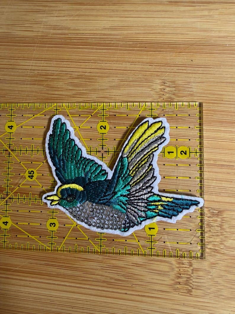 May include: An embroidered bird patch featuring teal, yellow, and silver thread with a white border. The patch is positioned on a yellow ruler, showing a width of approximately 3.5 inches and a height of approximately 2.5 inches.