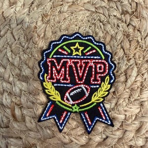 May include: A black embroidered patch shaped like a circular award ribbon with two tails. The patch features the red text MVP, a white football graphic, a yellow star, and yellow laurel branches, all outlined in bright green and white stitching.