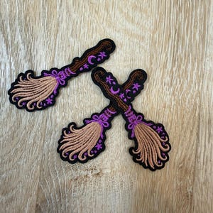 May include: Three embroidered patches shaped like broomsticks with brown handles and tan bristles. Each patch features purple stitching details including a crescent moon, small stars, and a bow tied around the base of the bristles, all outlined in black.