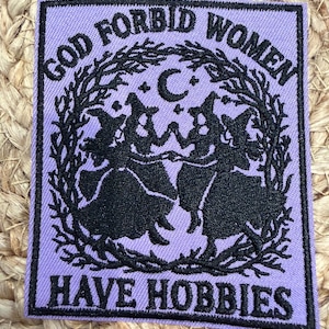 May include: A rectangular embroidered patch with a light purple background and black stitching. It features a central circular wreath of branches enclosing silhouettes of three figures in pointed hats, with the text GOD FORBID WOMEN HAVE HOBBIES.